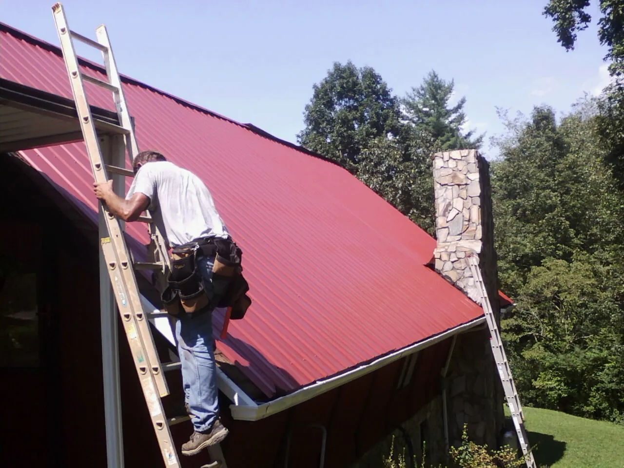 Roofer on a ladder beside a red metal roof and stone chimney — Soffit & Fascia Repair by Roofing Brothers in Indianola